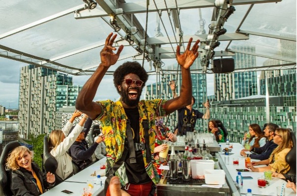 Man in a colourful mainly yellow shirt laughing with his hands in the air surrounded by people sitting in chairs around a white table suspended high in the air