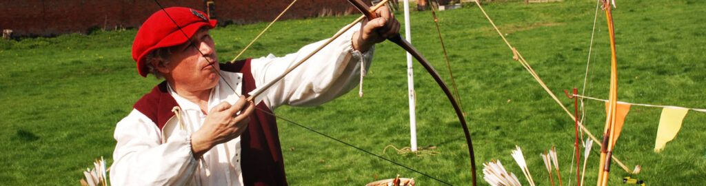a man, standing on grass, in a red beret leather waistcoat and white shire aiming an arrow in a bow into the air.